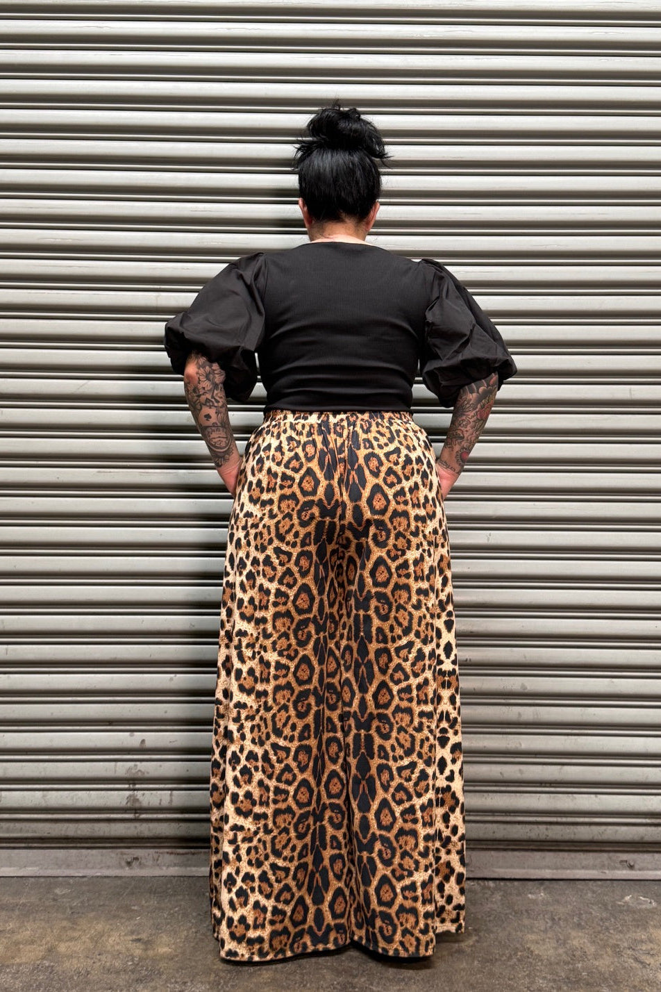 A person viewed from above wearing a black top and a leopard-print skirt, standing in front of a corrugated metal wall.