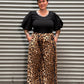 A woman wearing a black short-sleeve top and a leopard-print skirt, standing in front of a gray corrugated metal wall.