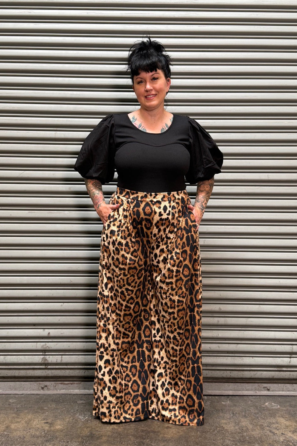 A woman wearing a black short-sleeve top and a leopard-print skirt, standing in front of a gray corrugated metal wall.