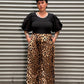 Woman with black hair and tattoos wearing a puff sleeve top with leopard pants standing in front of a metal back drop.