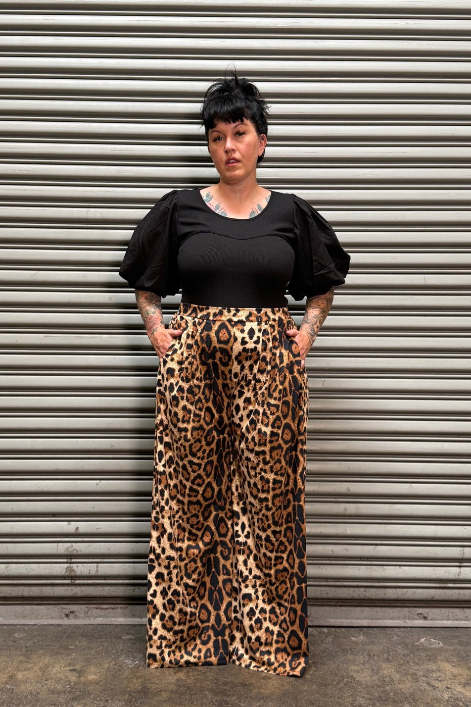 Woman with black hair and tattoos wearing a puff sleeve top with leopard pants standing in front of a metal back drop.
