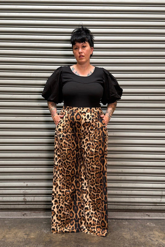 Woman with black hair and tattoos wearing a puff sleeve top with leopard pants standing in front of a metal back drop.