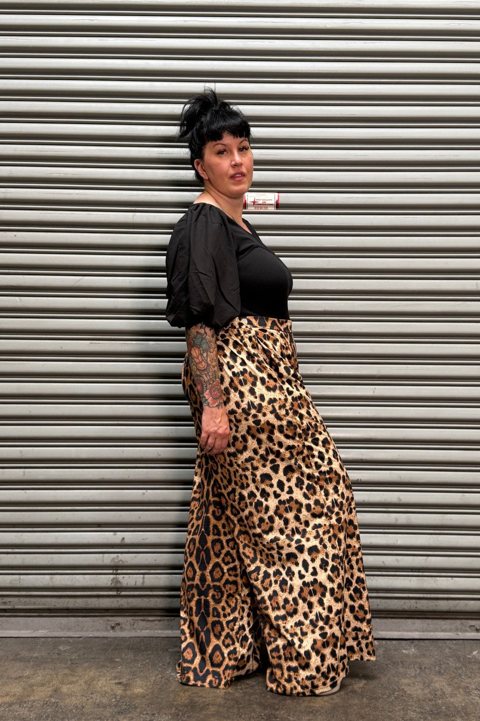 A person wearing leopard-print pants and a black top stands in front of a gray corrugated metal shutter.