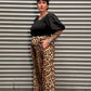 Woman with black hair and tattoos wearing a puff sleeve top with leopard pants standing to the side by a metal back drop.
