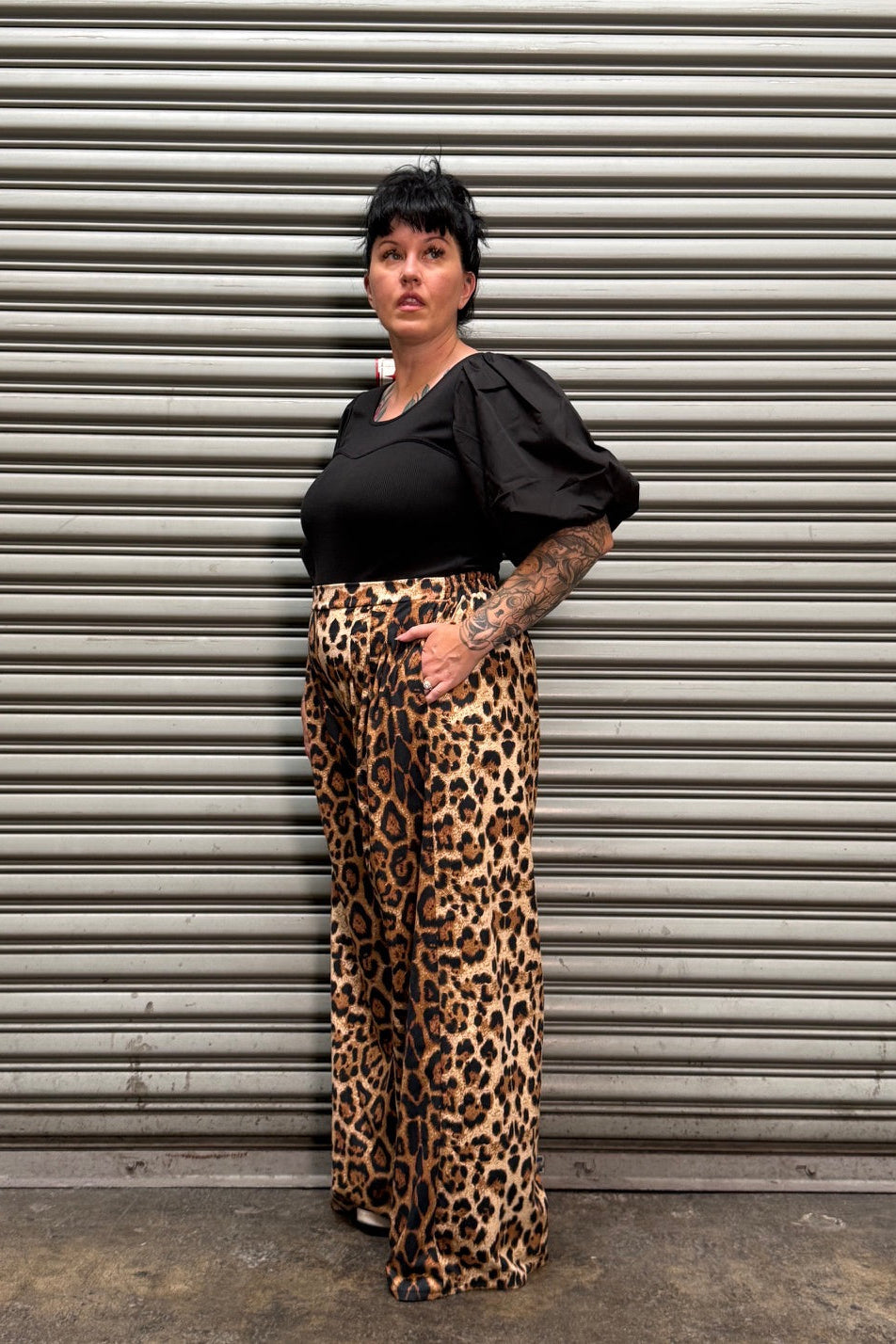 Woman with black hair and tattoos wearing a puff sleeve top with leopard pants standing to the side by a metal back drop.