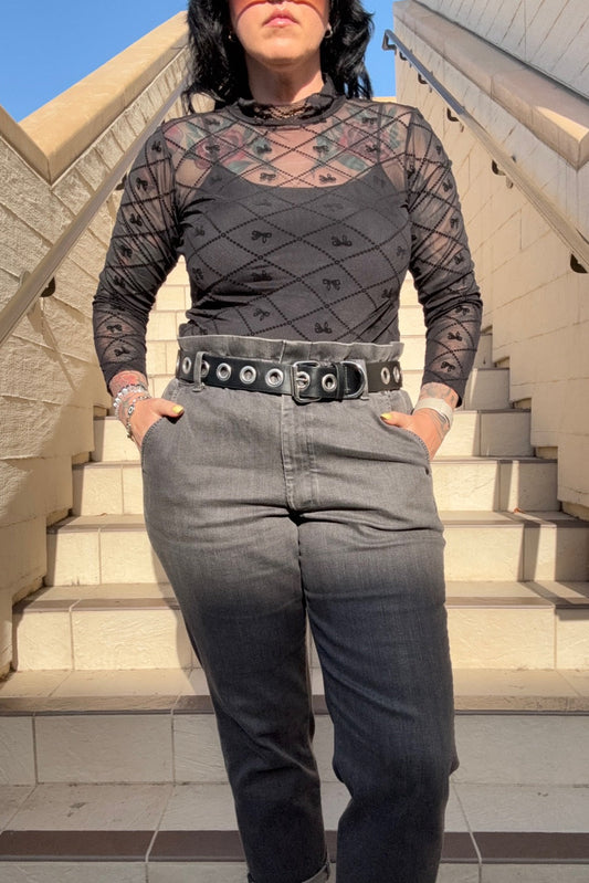 A person wearing a long sleeve black mesh top with a bow pattern, paired with gray trousers, standing on steps.