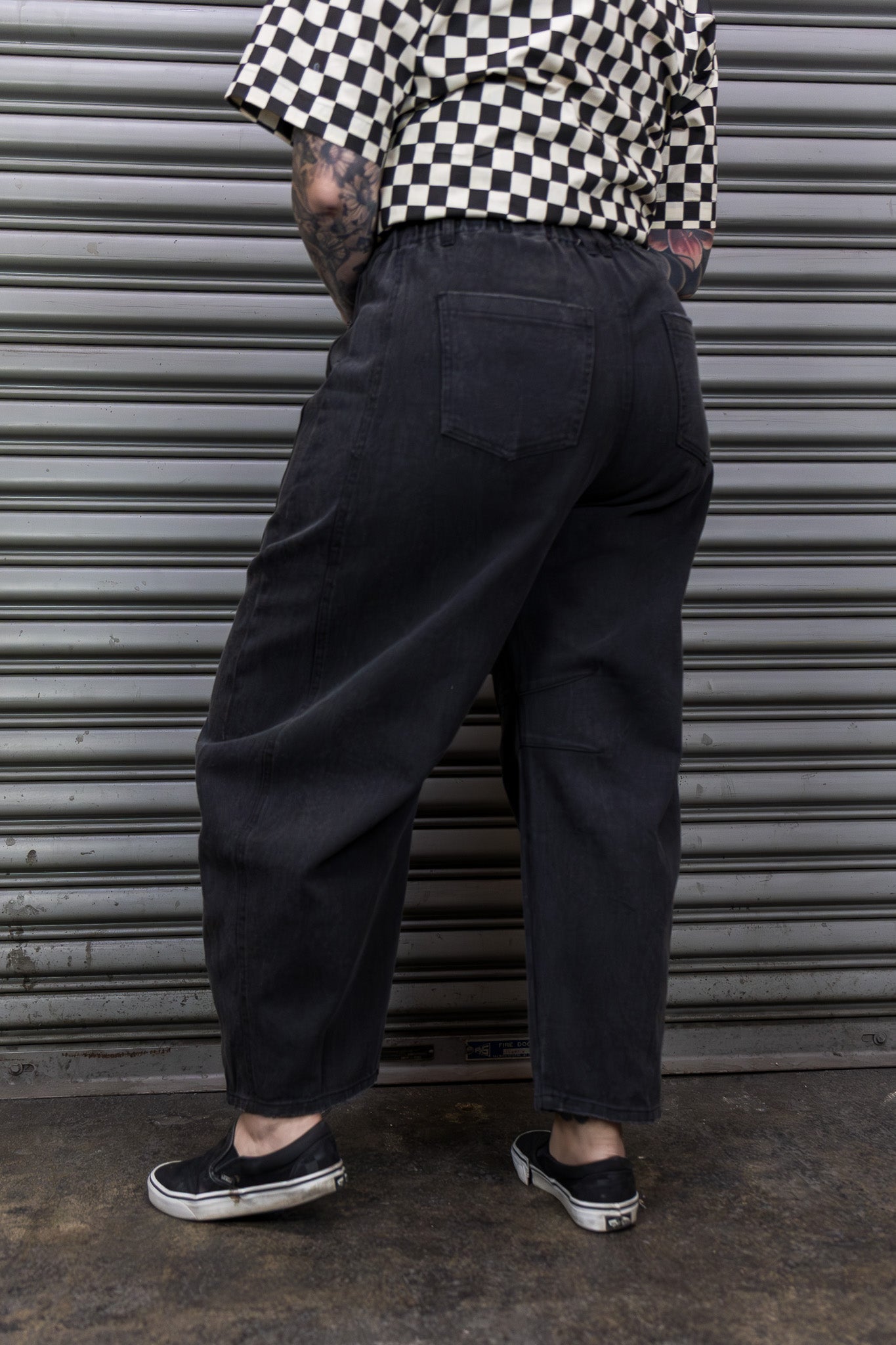 Person wearing black pants and a checkered shirt standing in front of a gray metal shutter.
