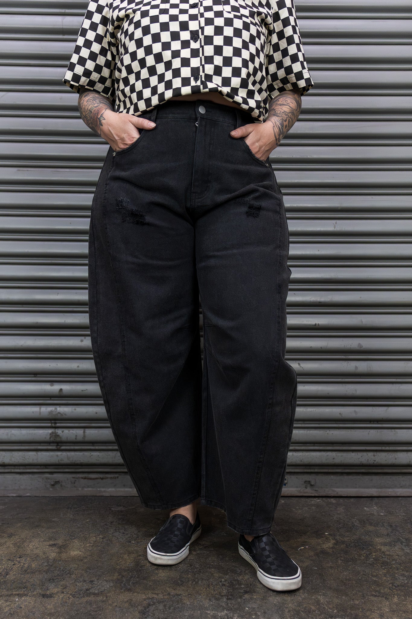 Person wearing a checkered shirt and black pants standing in front of a metallic shutter.
