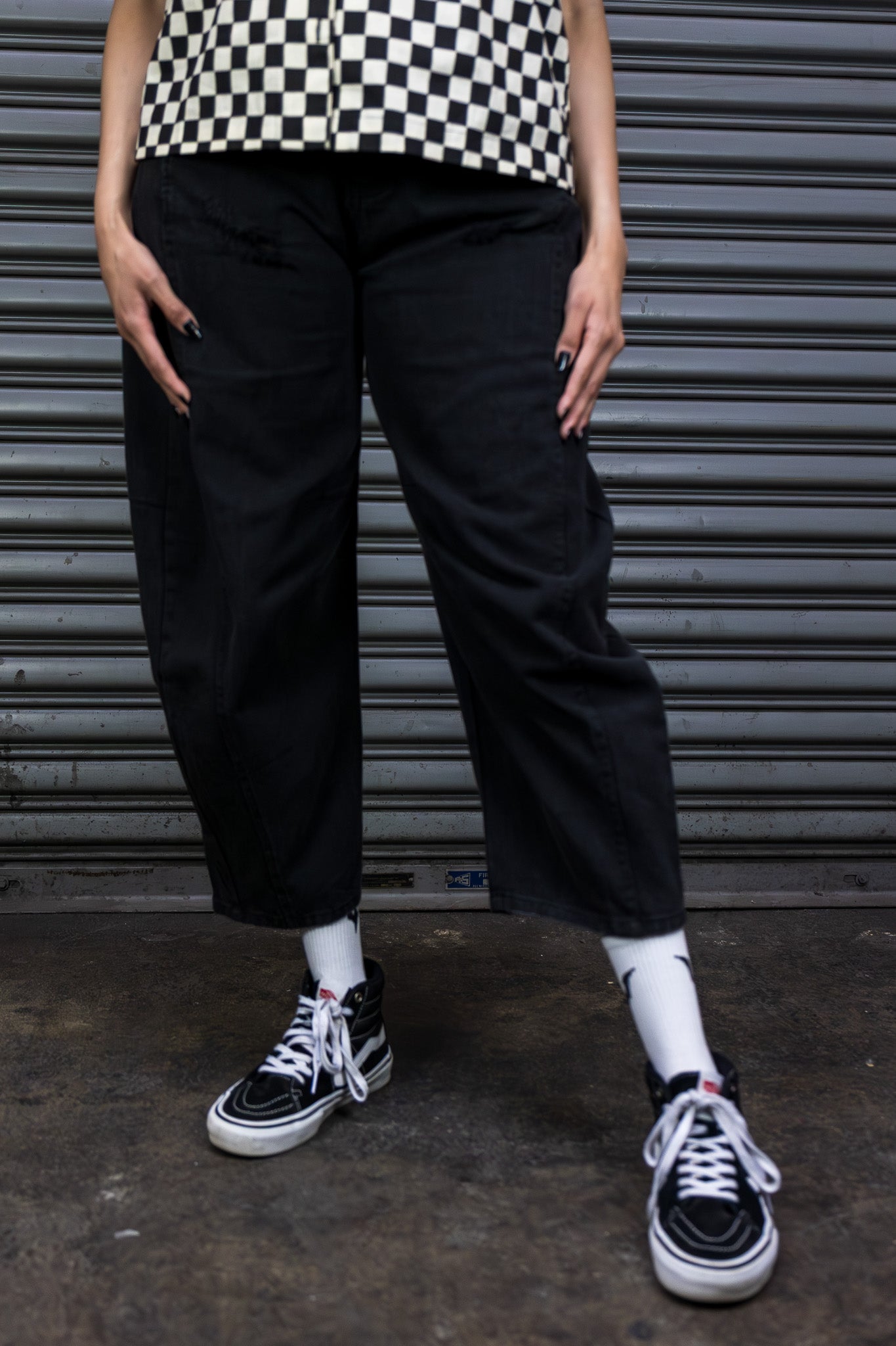 Person wearing a checkered shirt, black pants, and black sneakers against a metallic shutter background