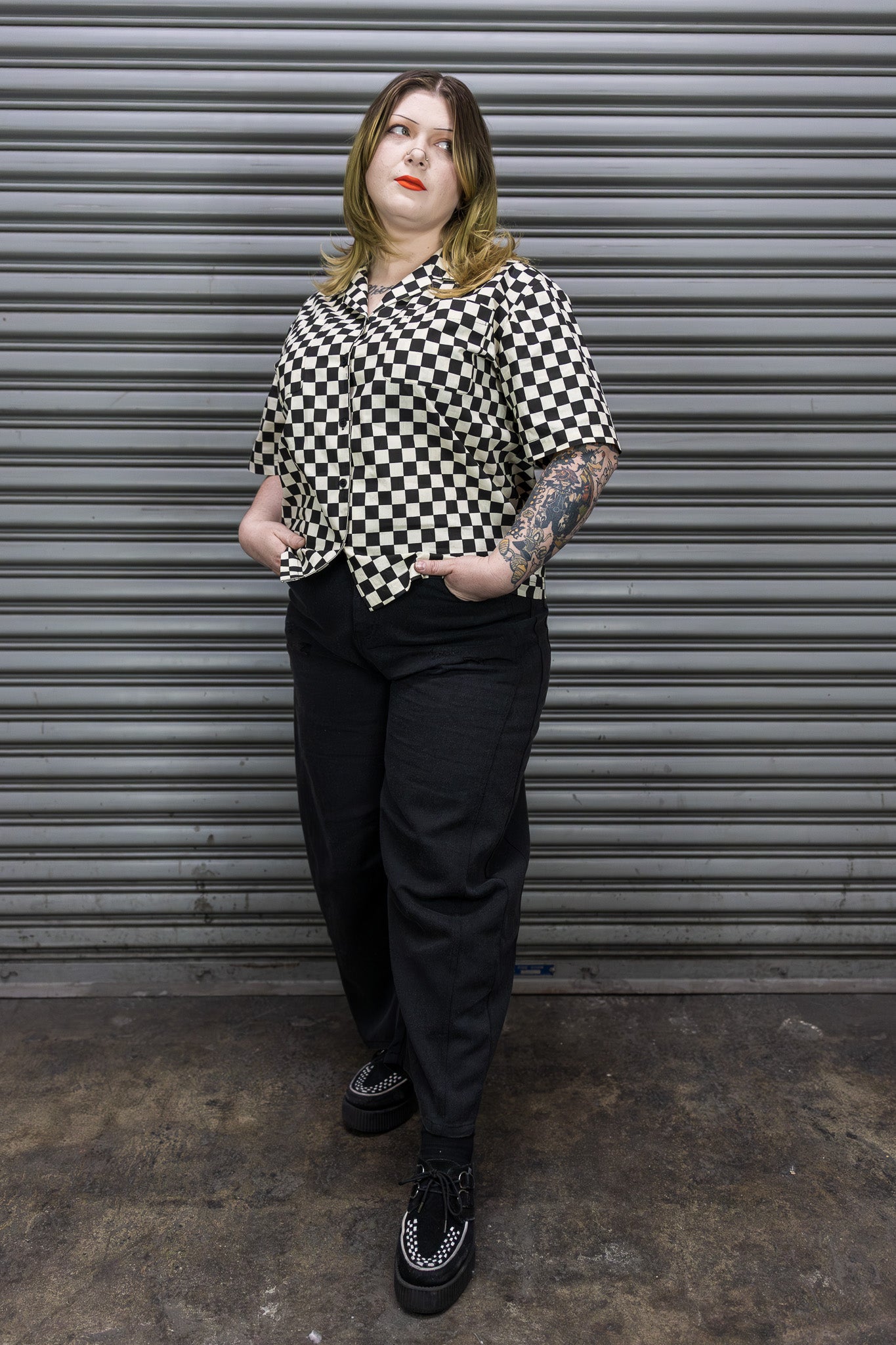 Person wearing a black and white checkered shirt and black pants standing in front of a metal shutter.