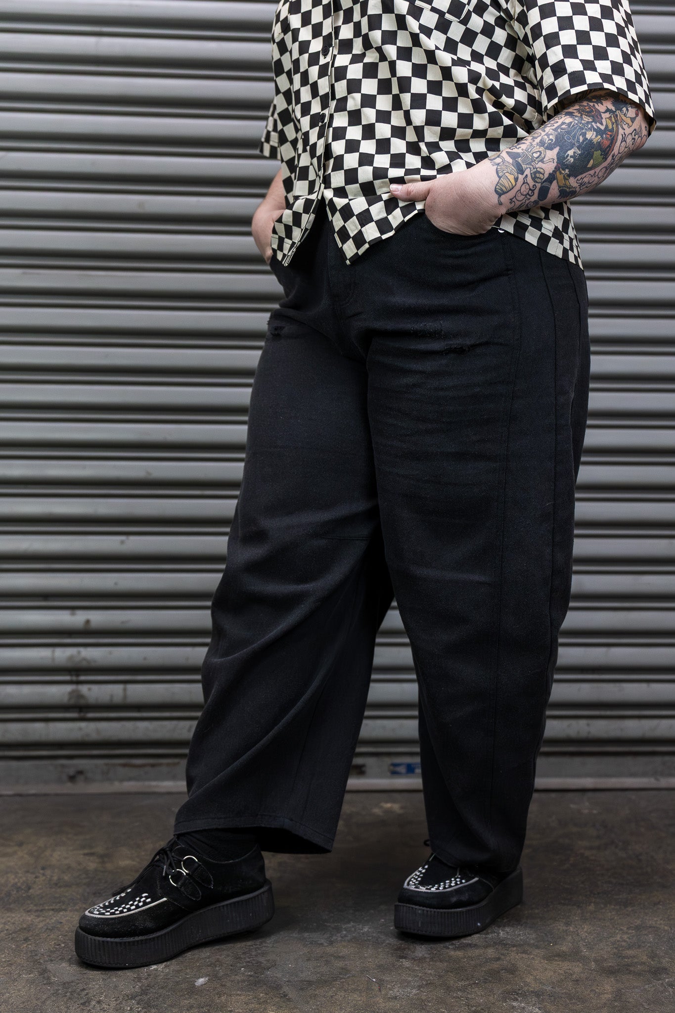 Person wearing a black and white checkered shirt, black pants, and black shoes against a metallic shutter background.