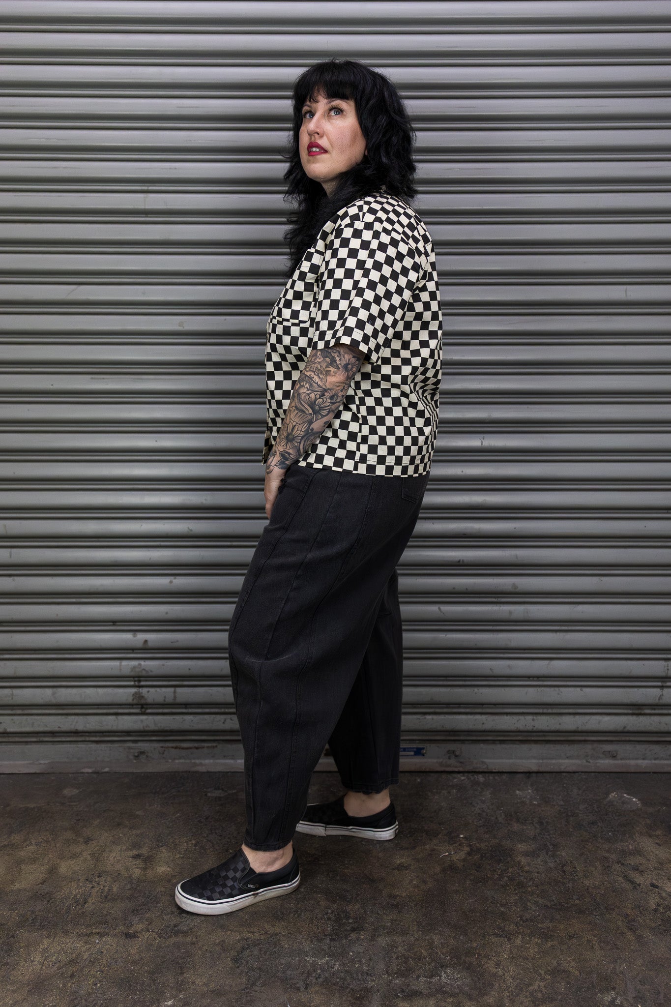 Person wearing a black and white checkered shirt with black pants standing in front of a metallic shutter.