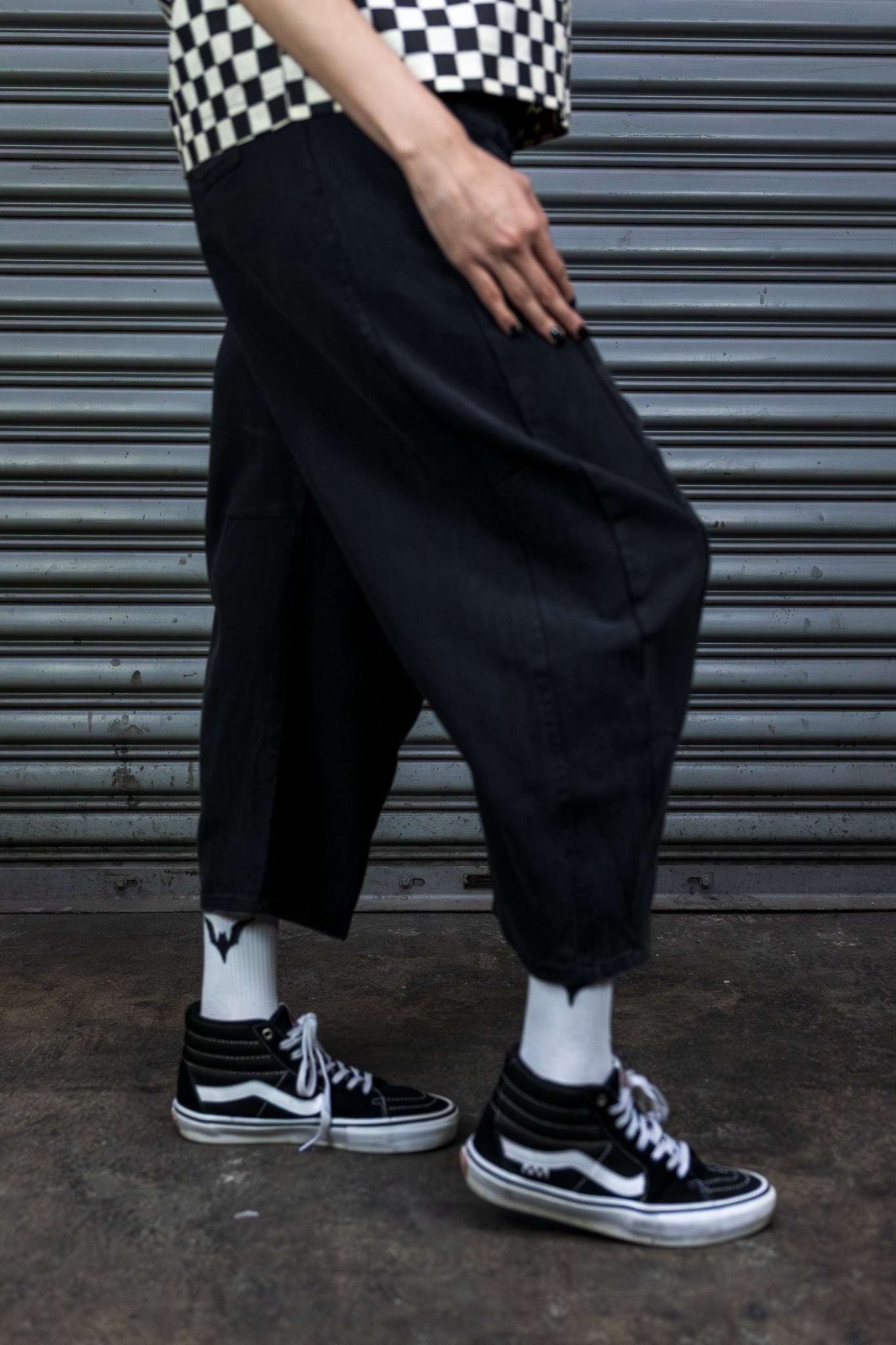Person wearing black pants and black and white sneakers in front of a metal shutter.