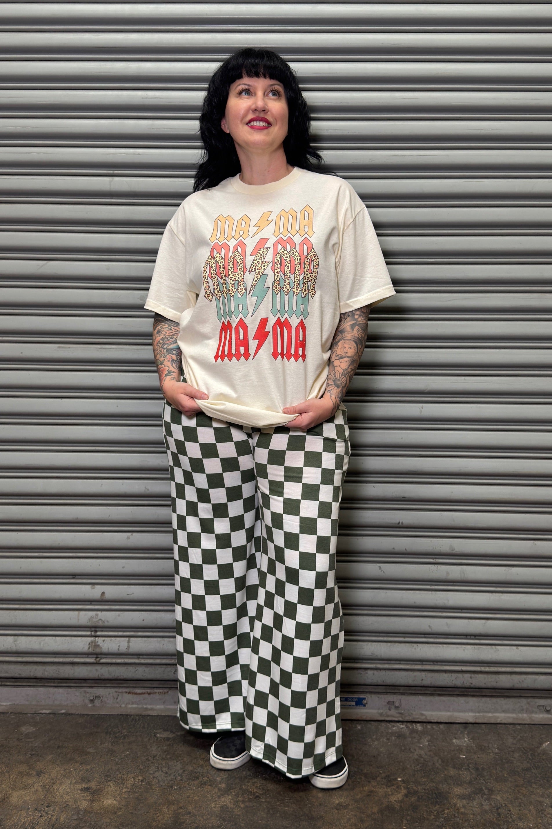 Person wearing a graphic t-shirt and checkered pants standing in front of a metal shutter.