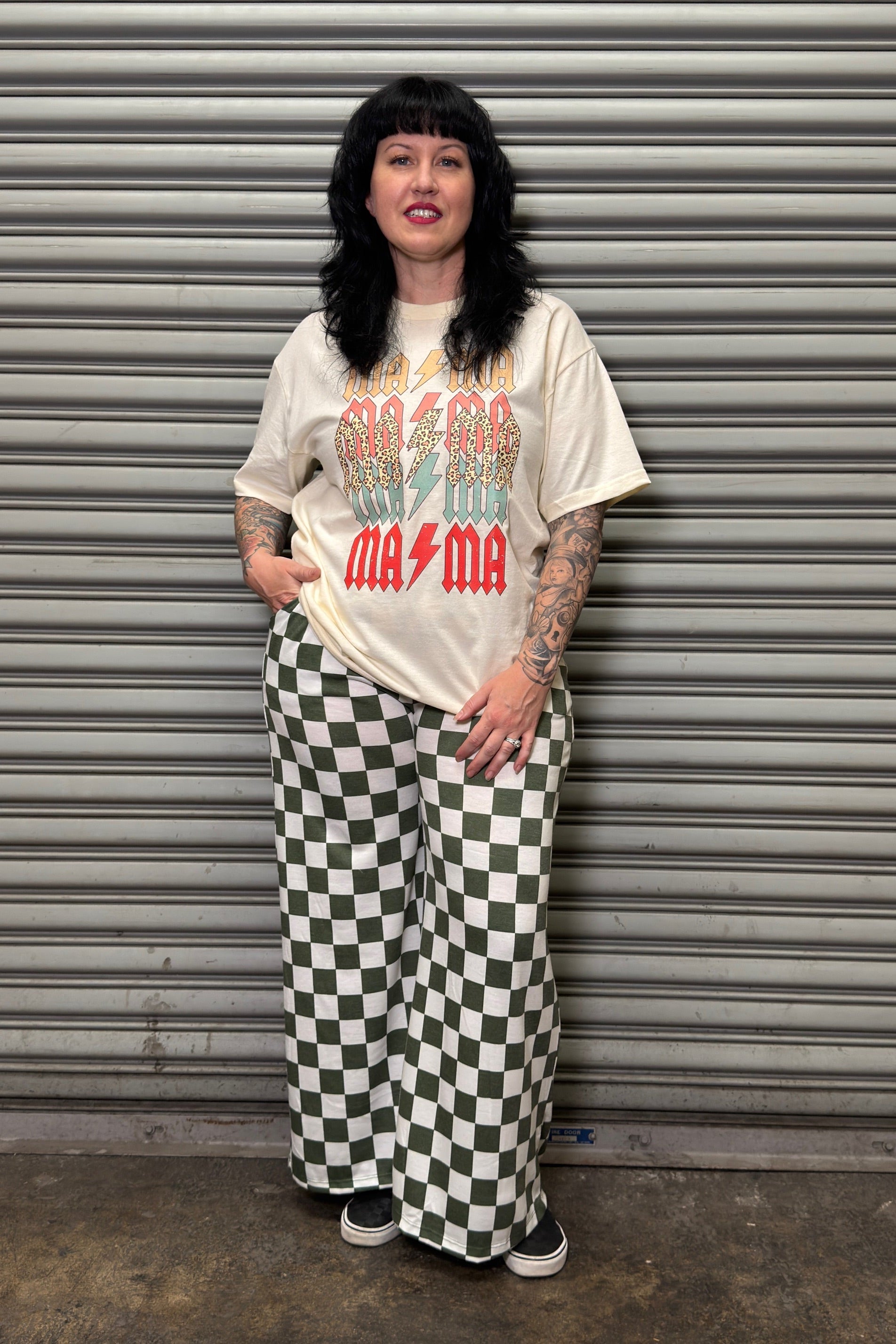 Person wearing a graphic t-shirt and checkered pants standing in front of a metal shutter.