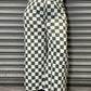 Person wearing a white t-shirt with colorful graphics and green and white checkered pants, leaning against a metallic shutter.