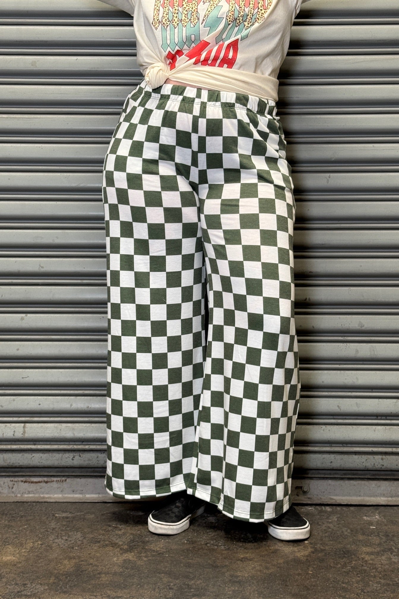 Person wearing a white t-shirt with colorful graphics and green and white checkered pants, leaning against a metallic shutter.