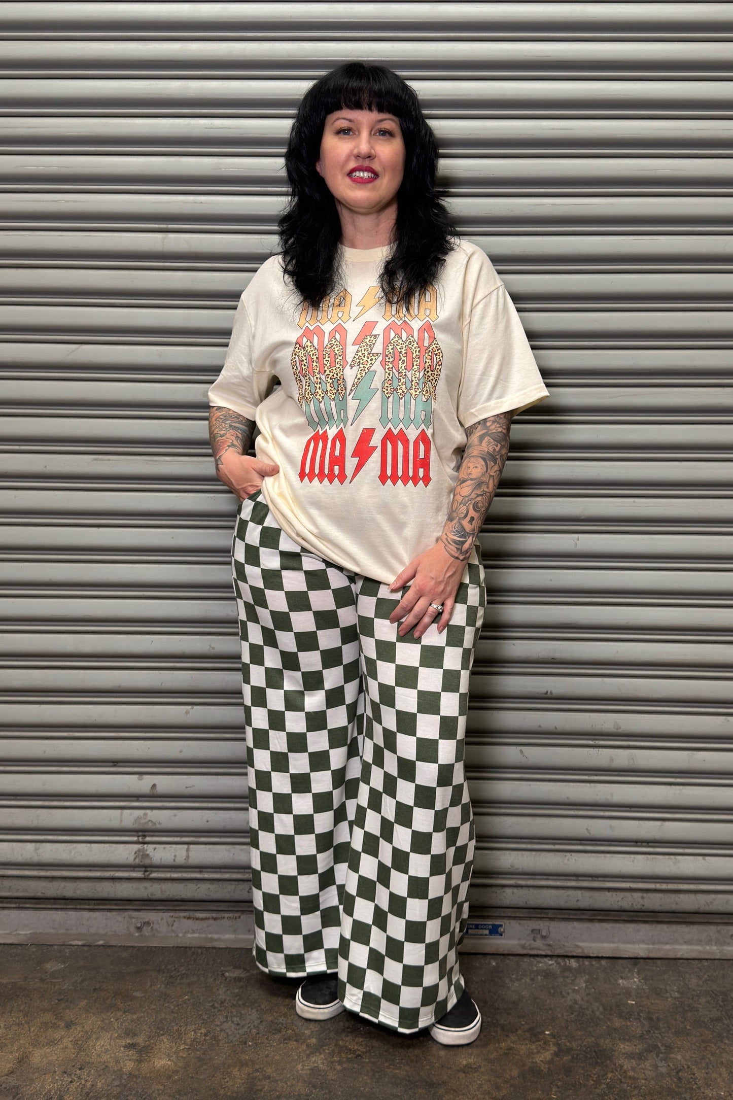 Person wearing a graphic t-shirt and checkered pants standing in front of a metal shutter.