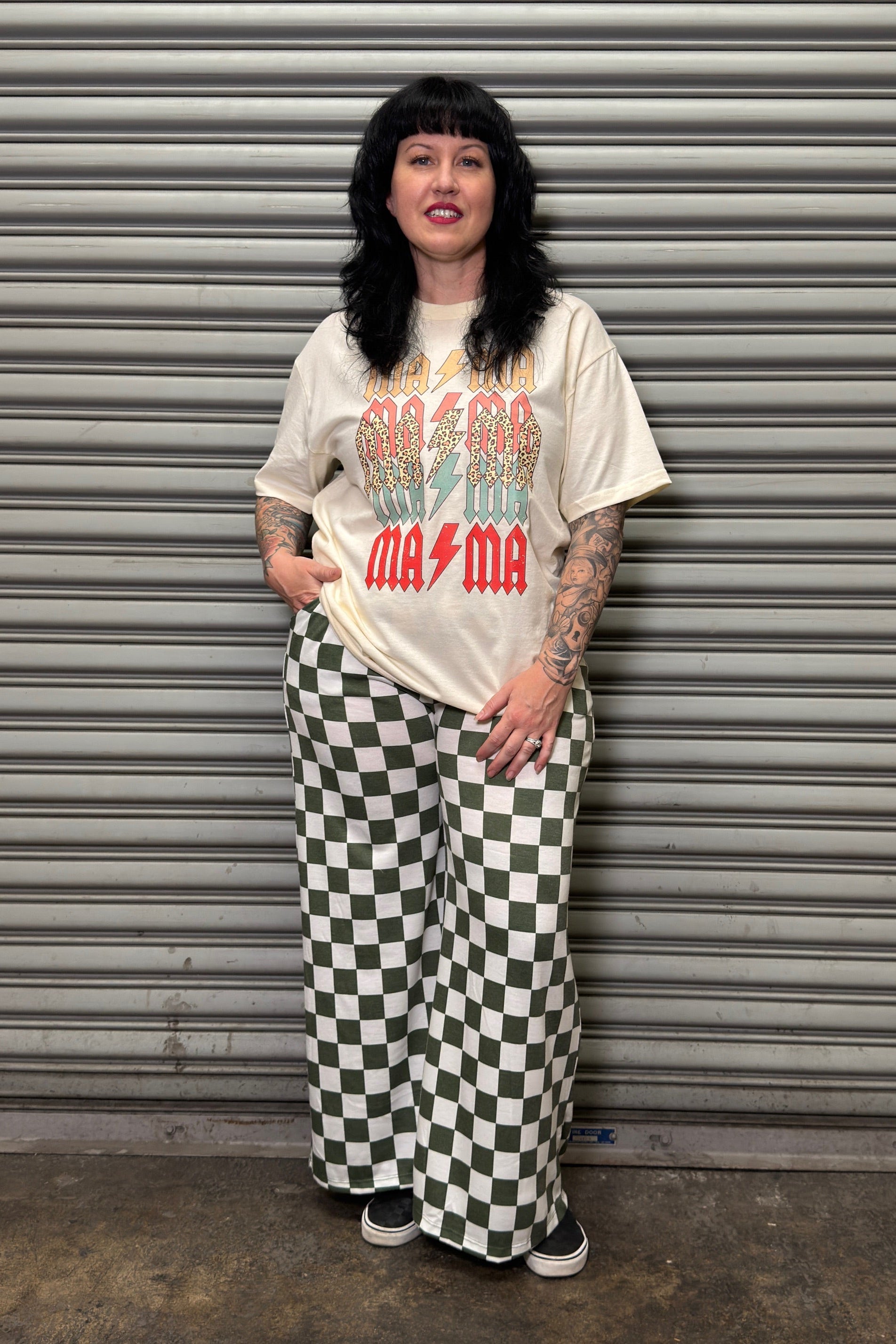 Person wearing a graphic t-shirt and checkered pants standing in front of a metal shutter.