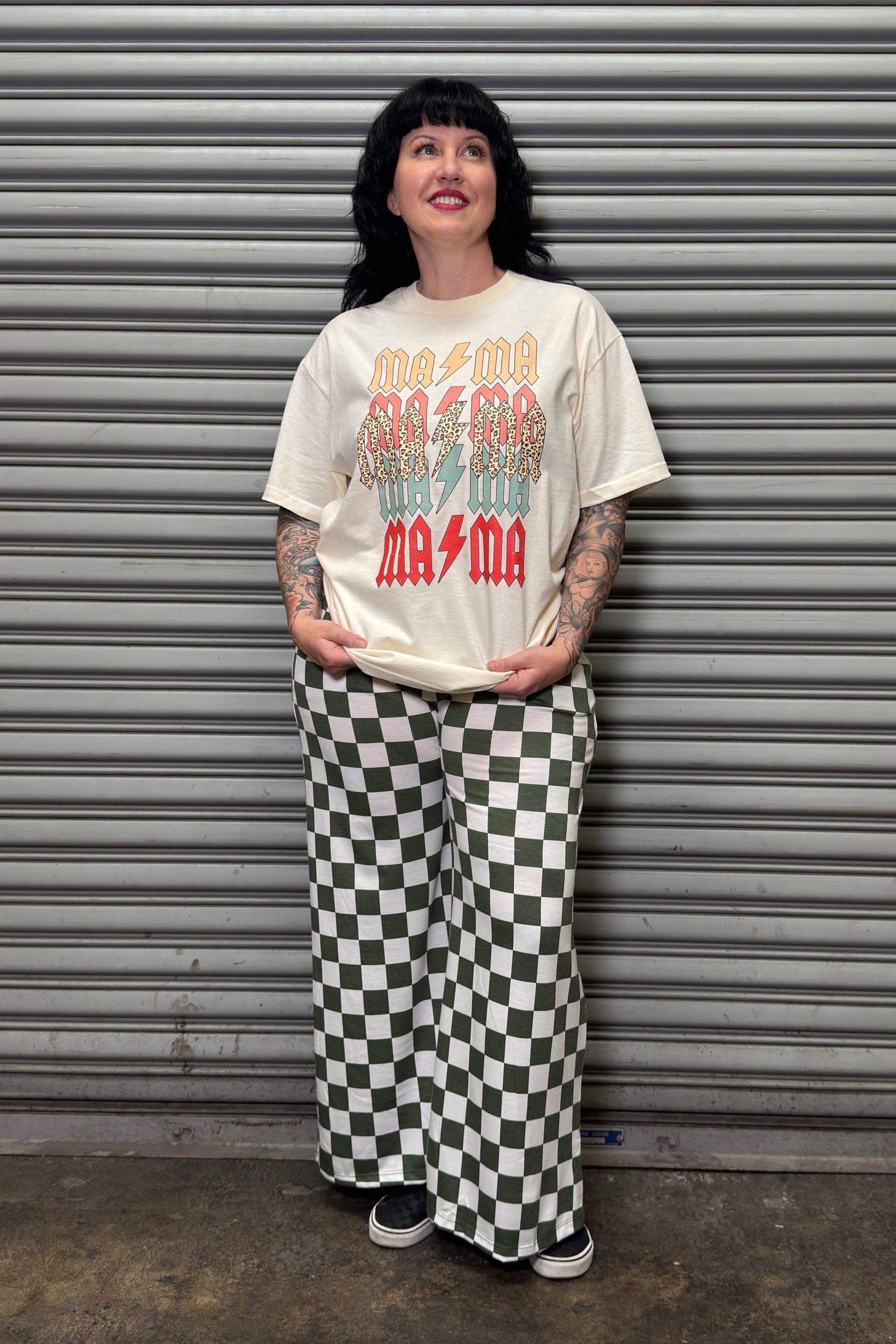 Person wearing a graphic t-shirt and checkered pants standing in front of a metal shutter.