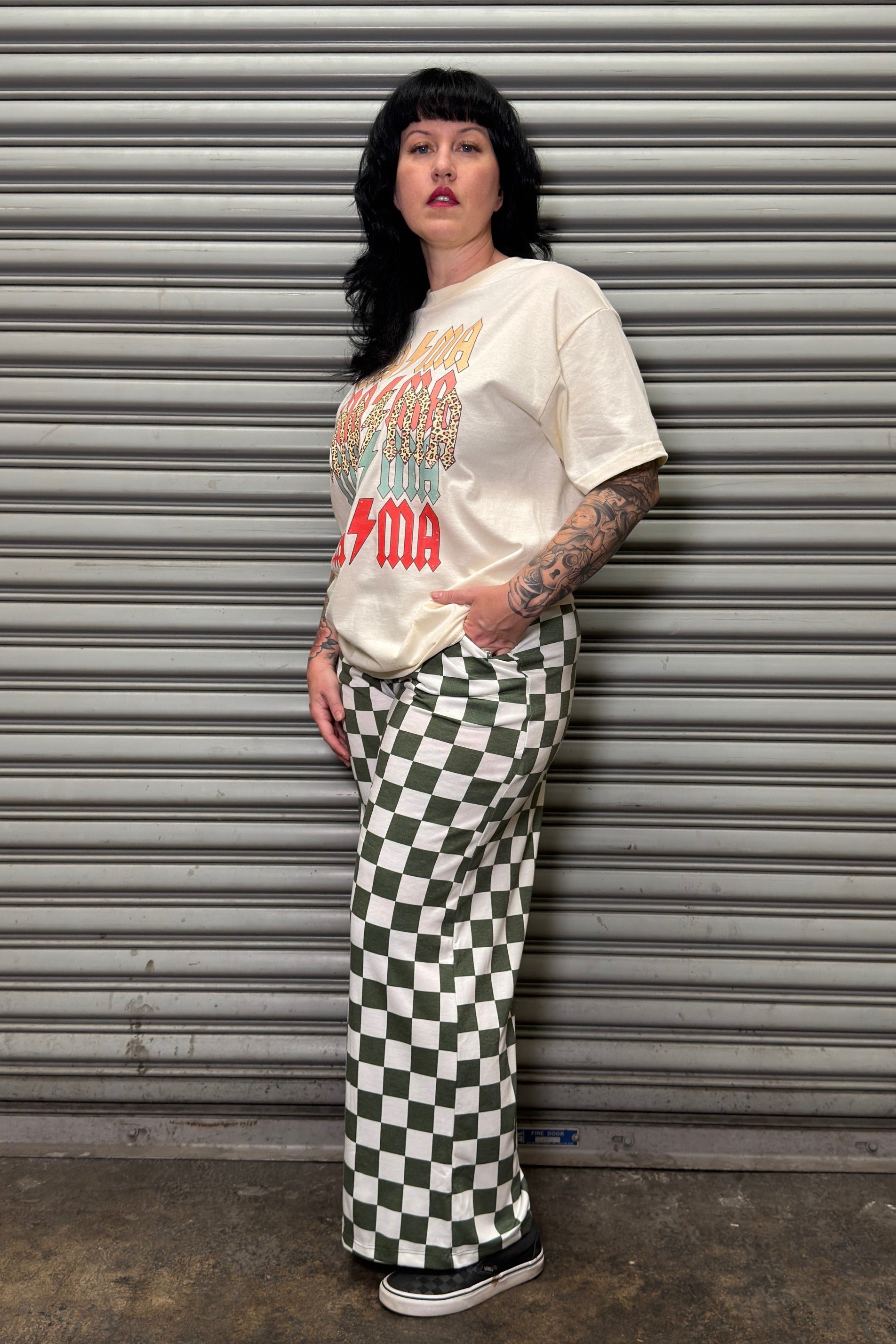Person wearing a graphic t-shirt and checkered pants leaning against a metal shutter.