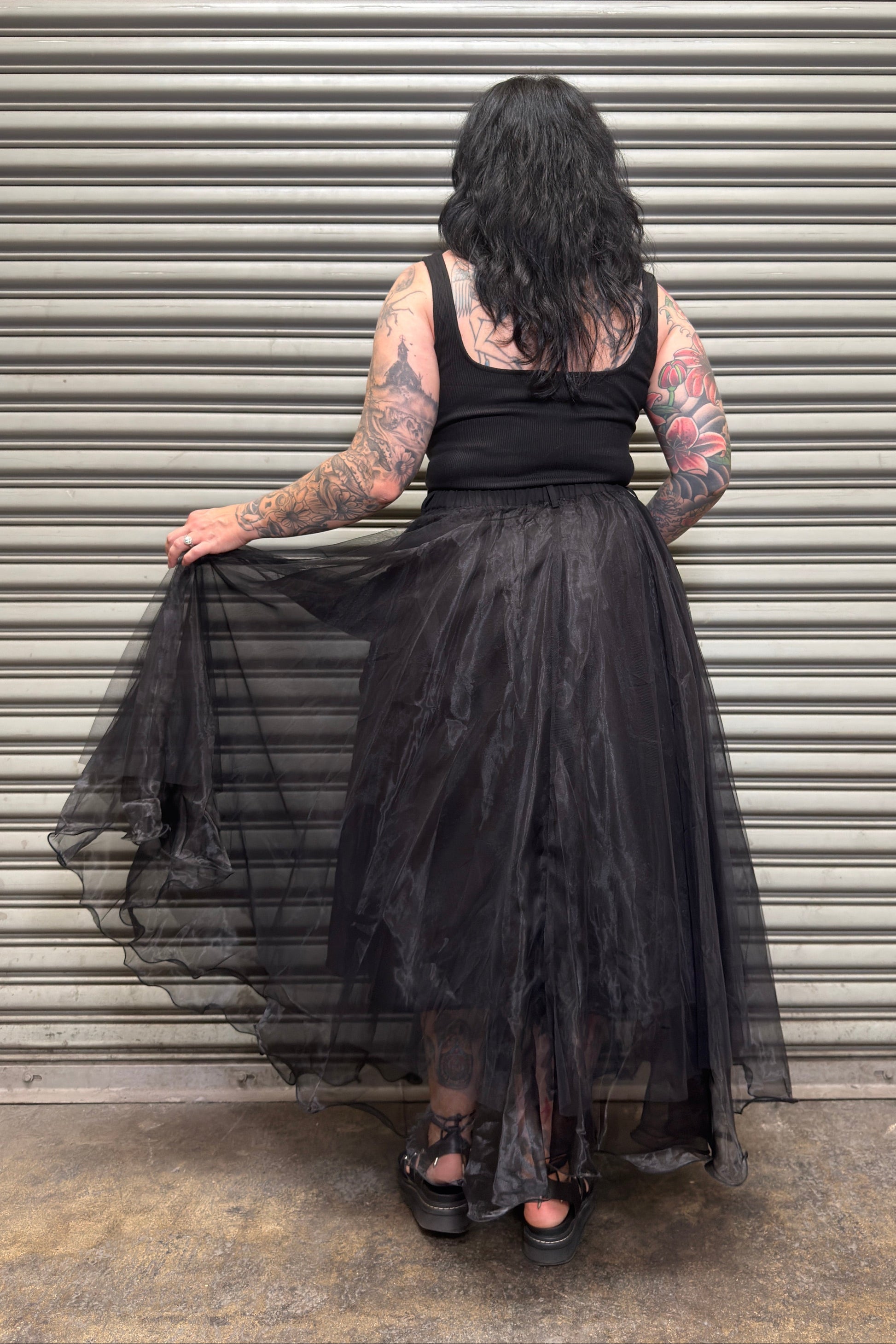 Person wearing a black dress with sheer overlay in front of a metallic shutter.
