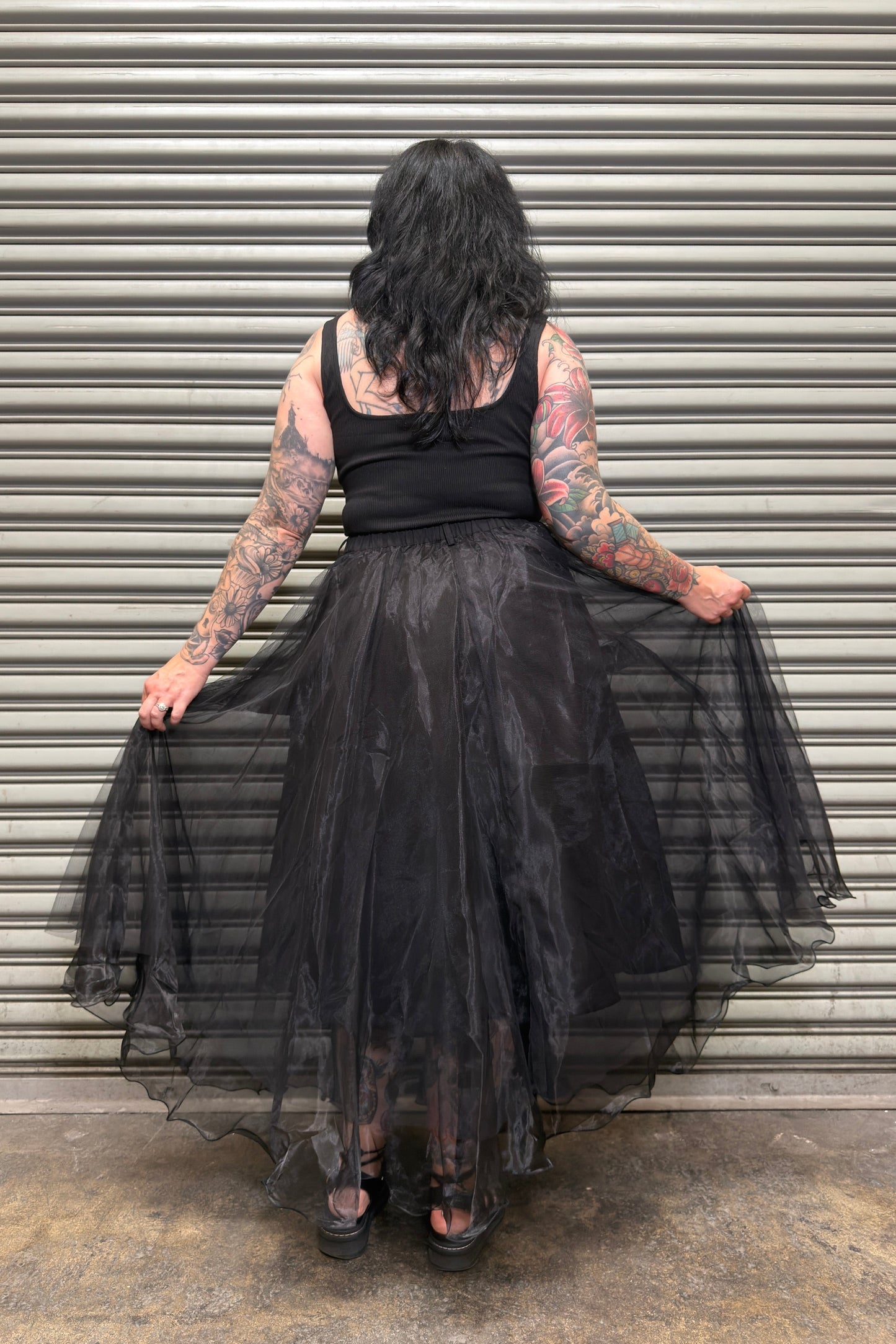 Person wearing a black dress with sheer overlay in front of a corrugated metal wall.