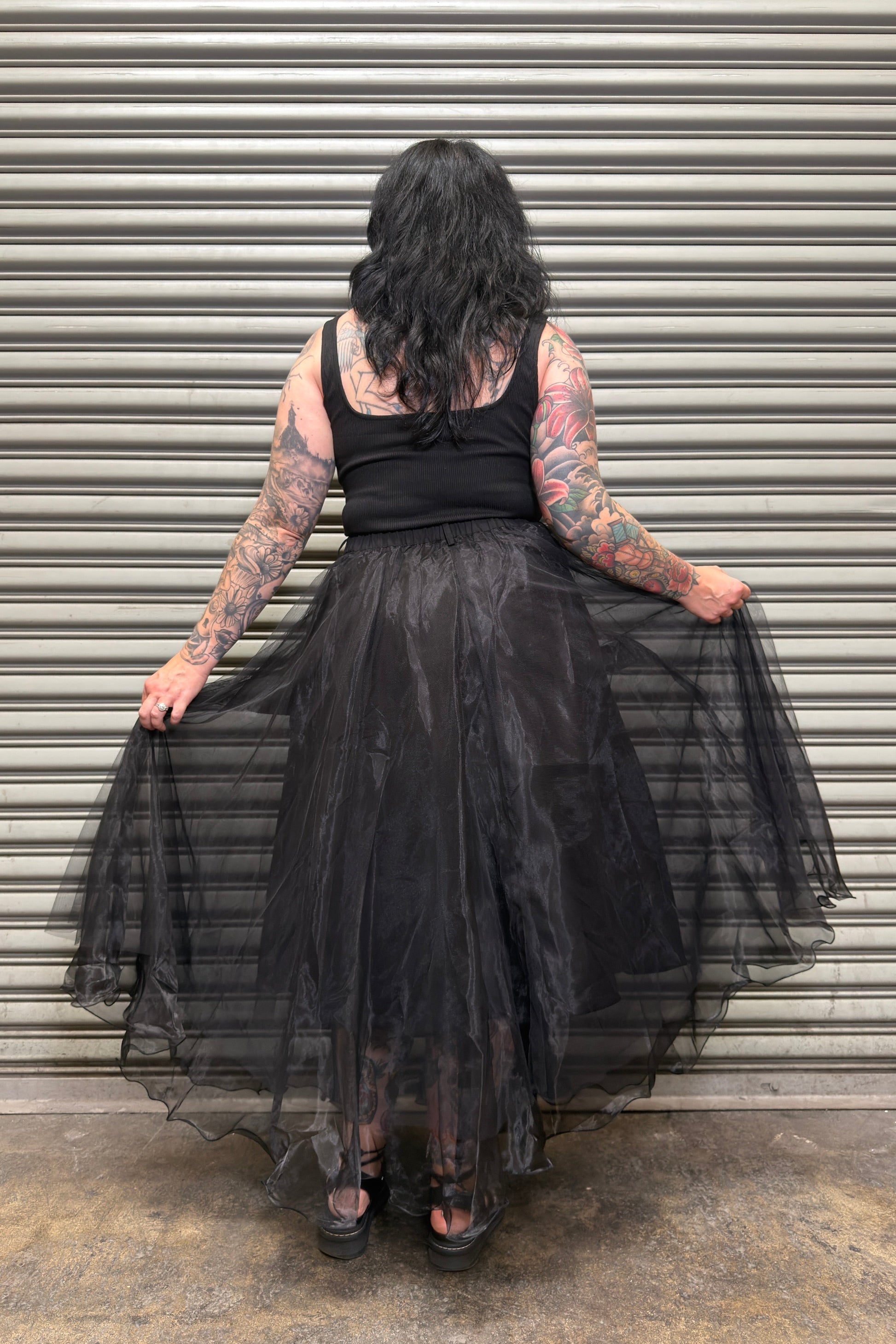 Person wearing a black dress with sheer overlay in front of a corrugated metal wall.