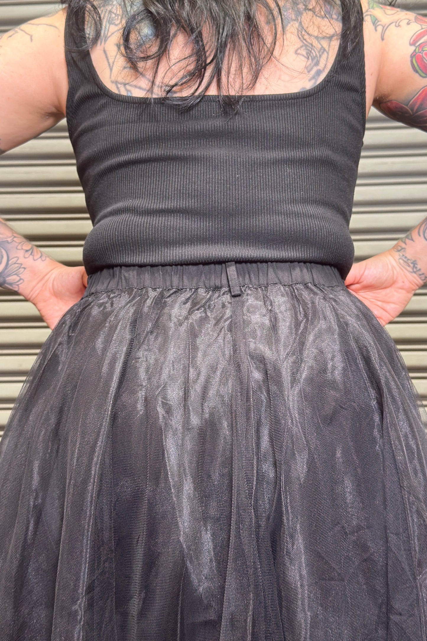 Person wearing a black tank top and tulle skirt with tattoos on arms and back.
