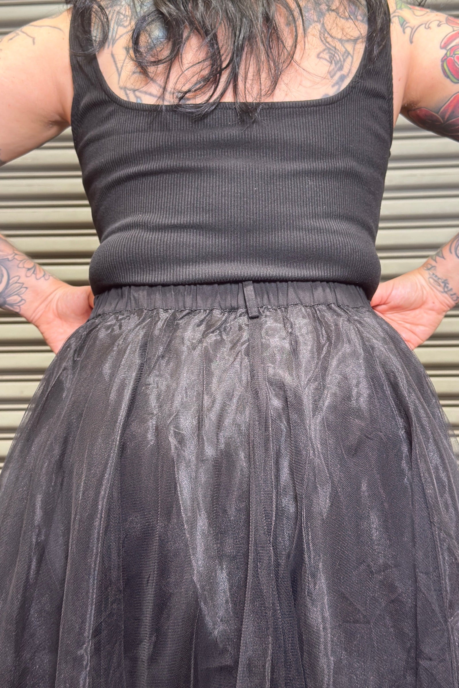 Person wearing a black tank top and tulle skirt with tattoos on arms and back.