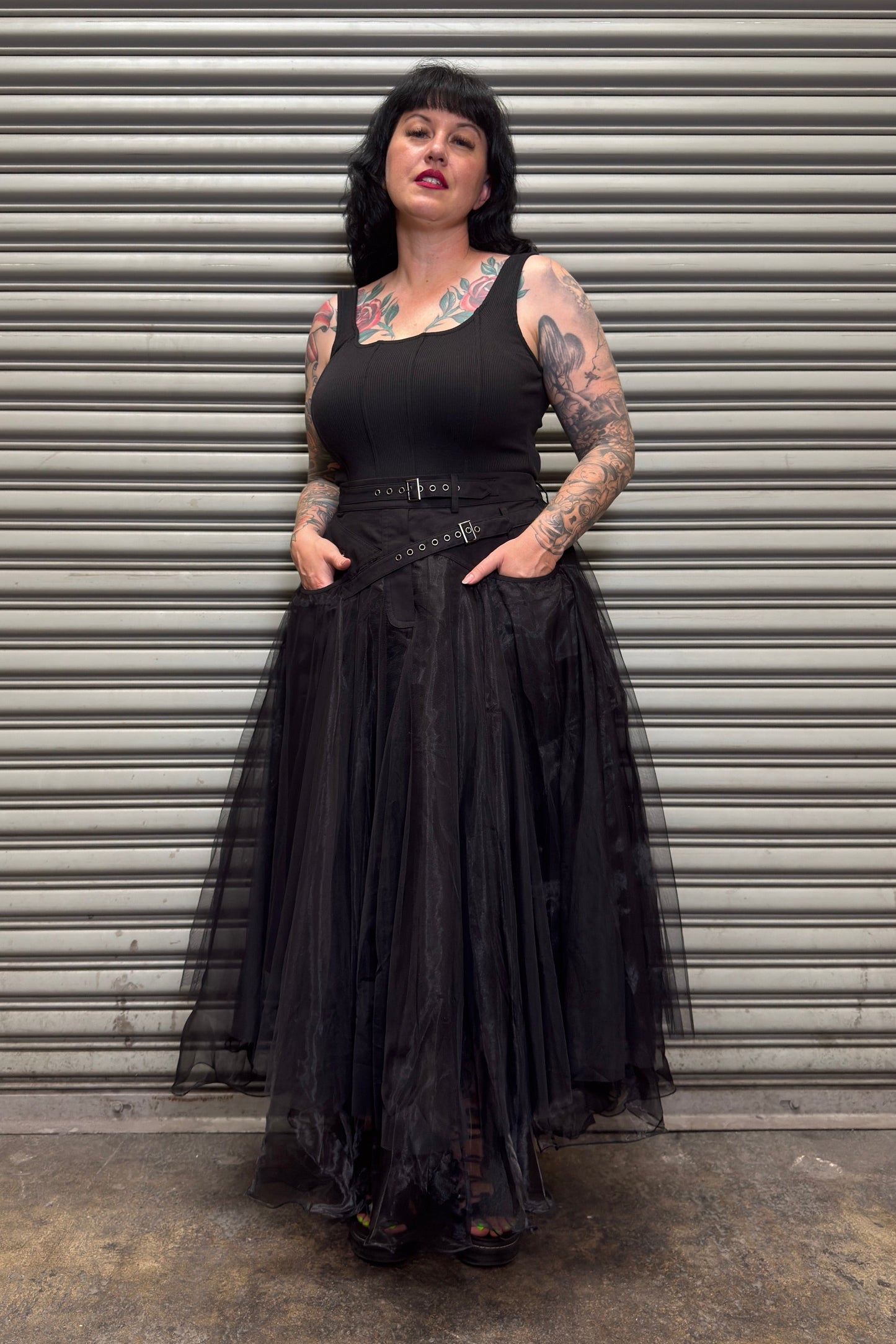 Person wearing a black dress with a tattooed arm standing in front of a metal shutter.