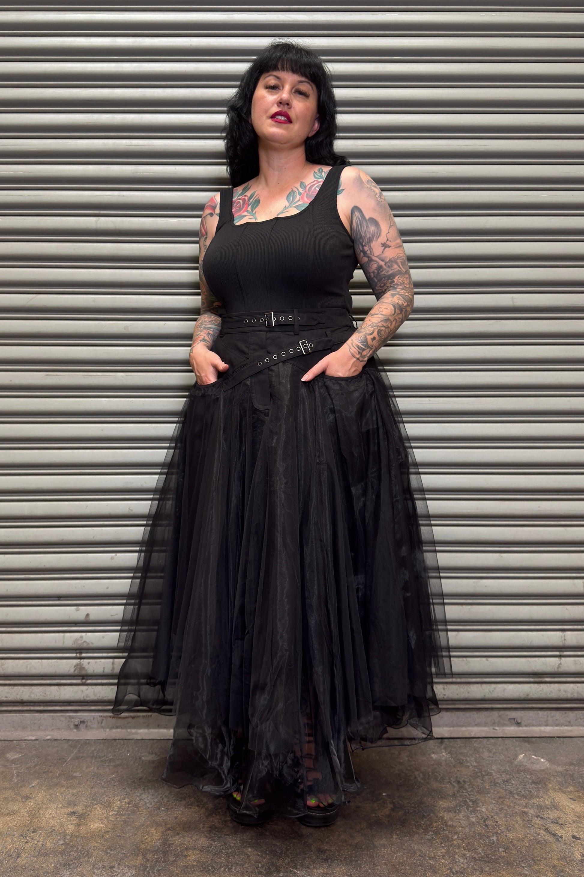 Person wearing a black dress with a tattooed arm standing in front of a metal shutter.