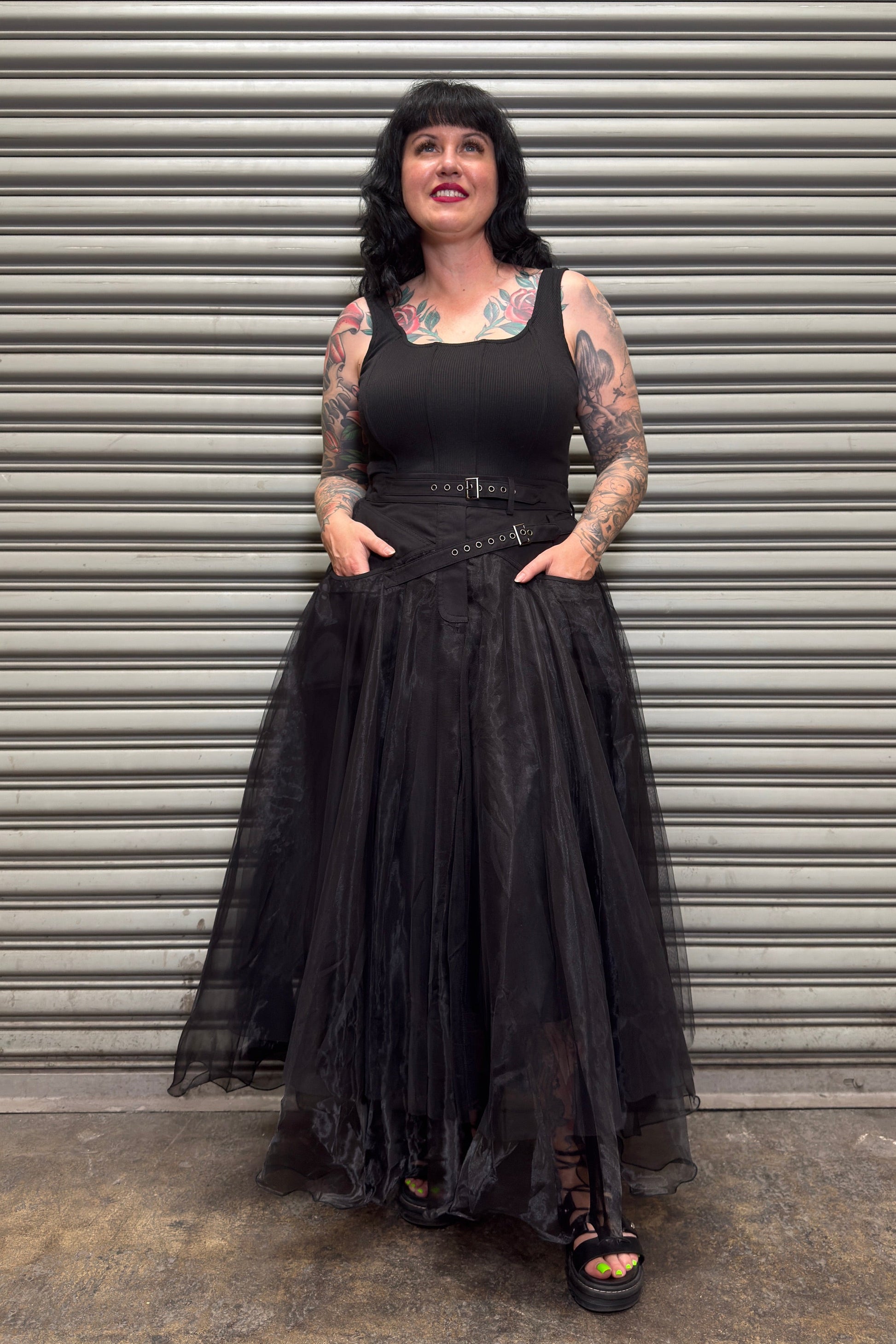 Woman wearing a black sleeveless top and long black skirt standing in front of a corrugated metal wall.