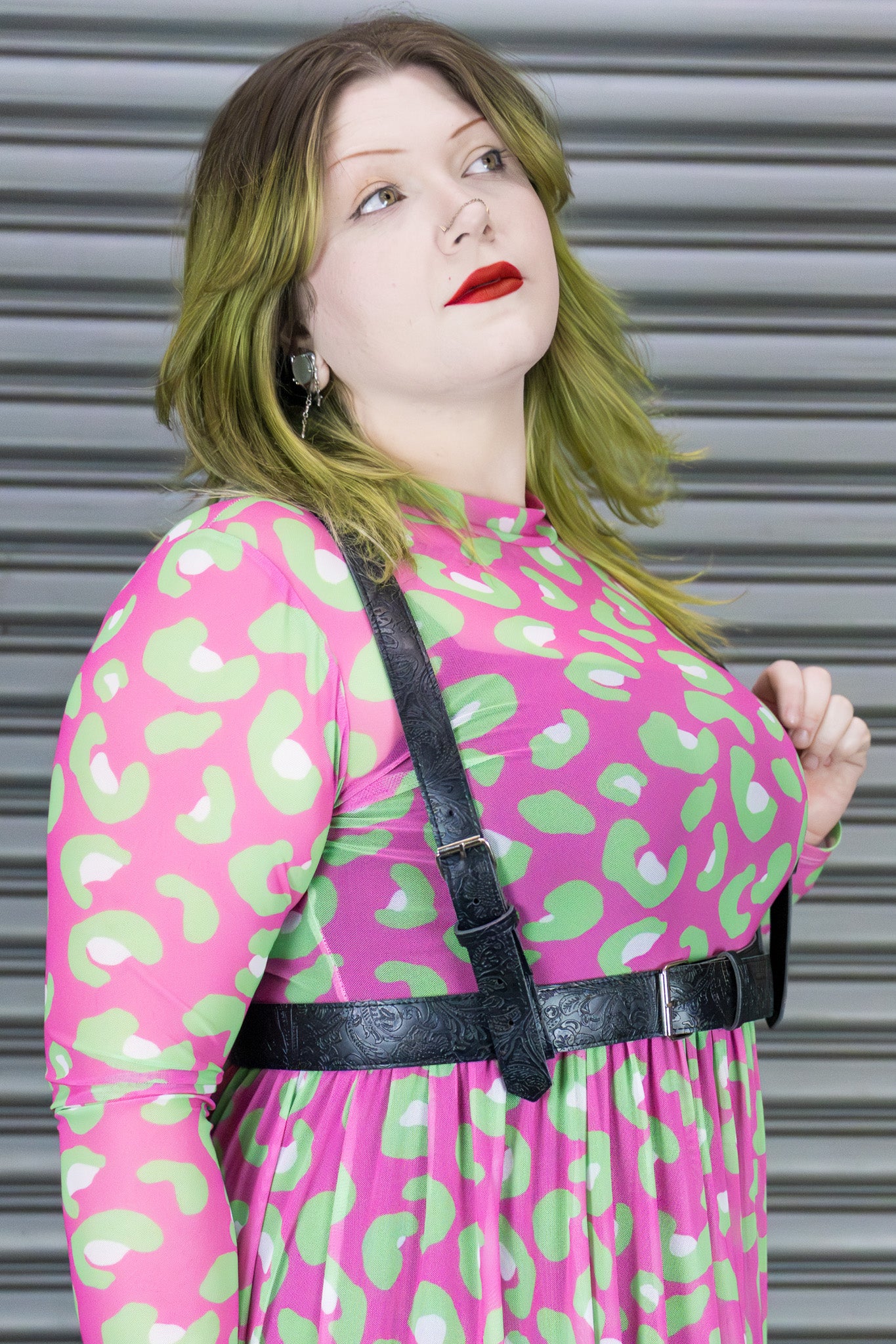 Woman wearing a pink dress with green leopard patterns and a black harness against a metallic background