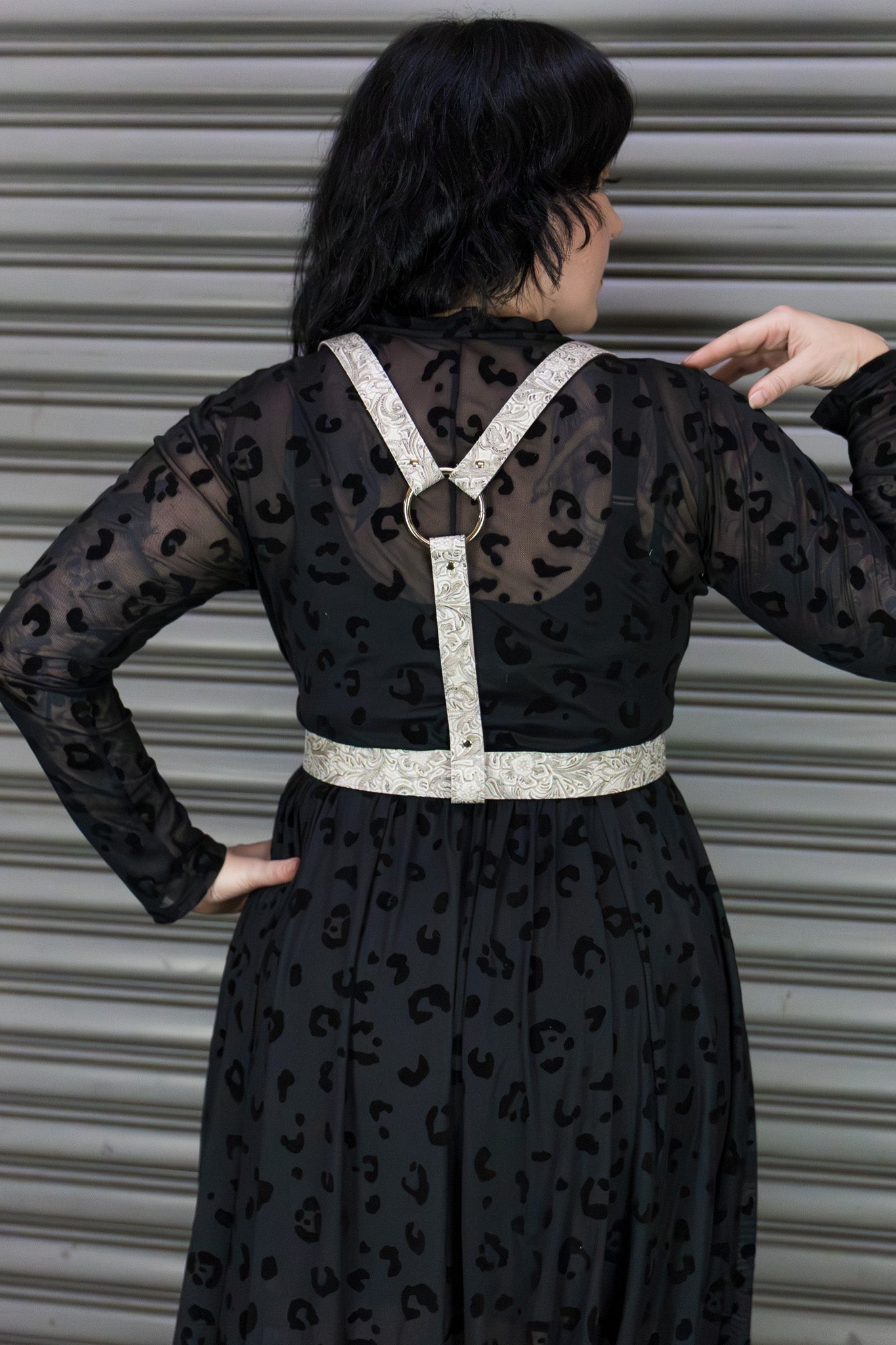 Person wearing a black dress with a leopard print pattern and a white harness against a metallic background.