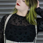 Person wearing a black dress with a sheer top and white harness against a metallic background.