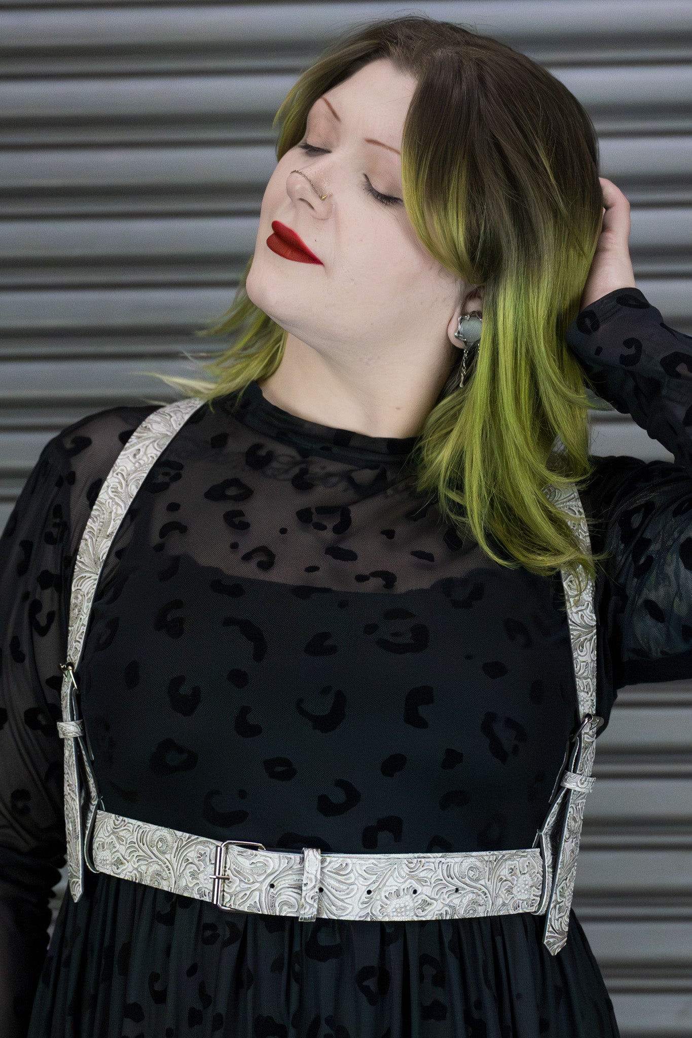 Person wearing a black dress with a sheer top and white harness against a metallic background.