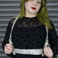 Person wearing a black dress with a sheer top and white harness against a metallic background