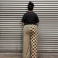 A person viewed from above wearing a black t-shirt and a long pleated skirt with a brown and beige checkered upper section and a beige-and-black striped lower section, standing against a corrugated metal wall.