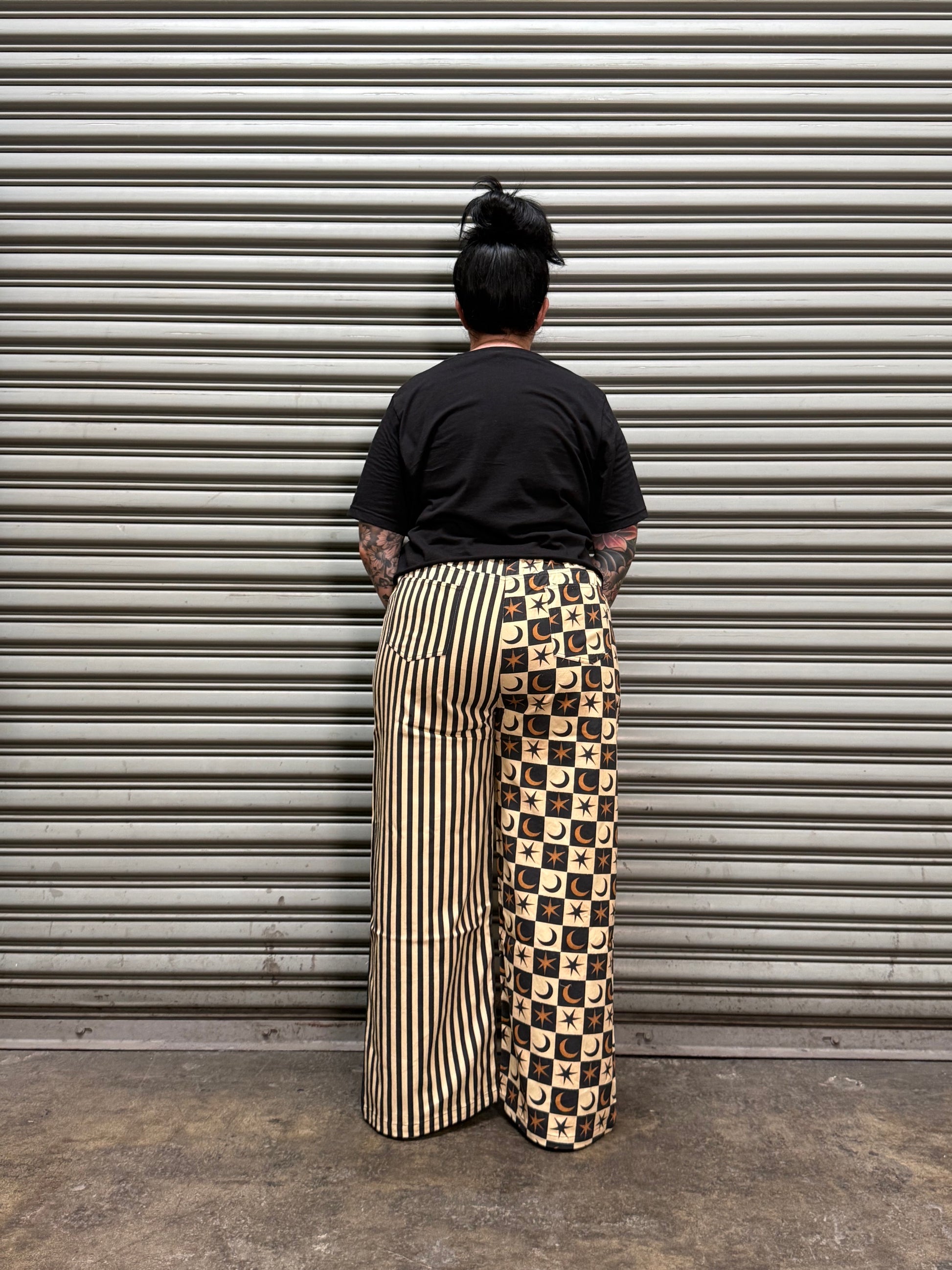 A person viewed from above wearing a black t-shirt and a long pleated skirt with a brown and beige checkered upper section and a beige-and-black striped lower section, standing against a corrugated metal wall.