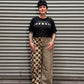A person wearing a black graphic t-shirt and beige-and-black patterned wide-leg pants, standing in front of a gray corrugated metal shutter.