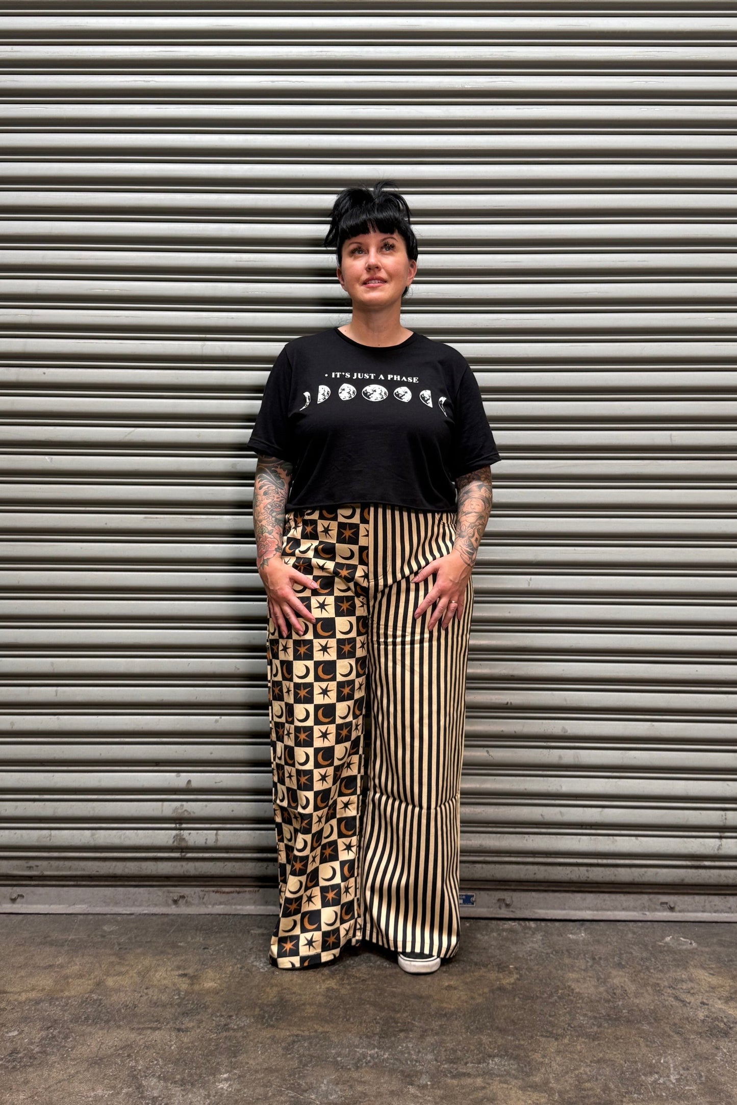 A person wearing a black graphic t-shirt and beige-and-black patterned wide-leg pants, standing in front of a gray corrugated metal shutter.