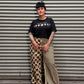 A person wearing a black graphic t-shirt and wide-leg pants with a beige and black striped pattern and a beige and black checkerboard pattern, standing in front of a corrugated metal shutter.