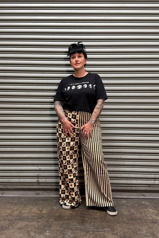 A person wearing a black graphic t-shirt and wide-leg pants with a beige and black striped pattern and a beige and black checkerboard pattern, standing in front of a corrugated metal shutter.