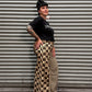 A person wearing a black t-shirt and a beige and black checkered maxi skirt, standing in front of a gray corrugated metal shutter.