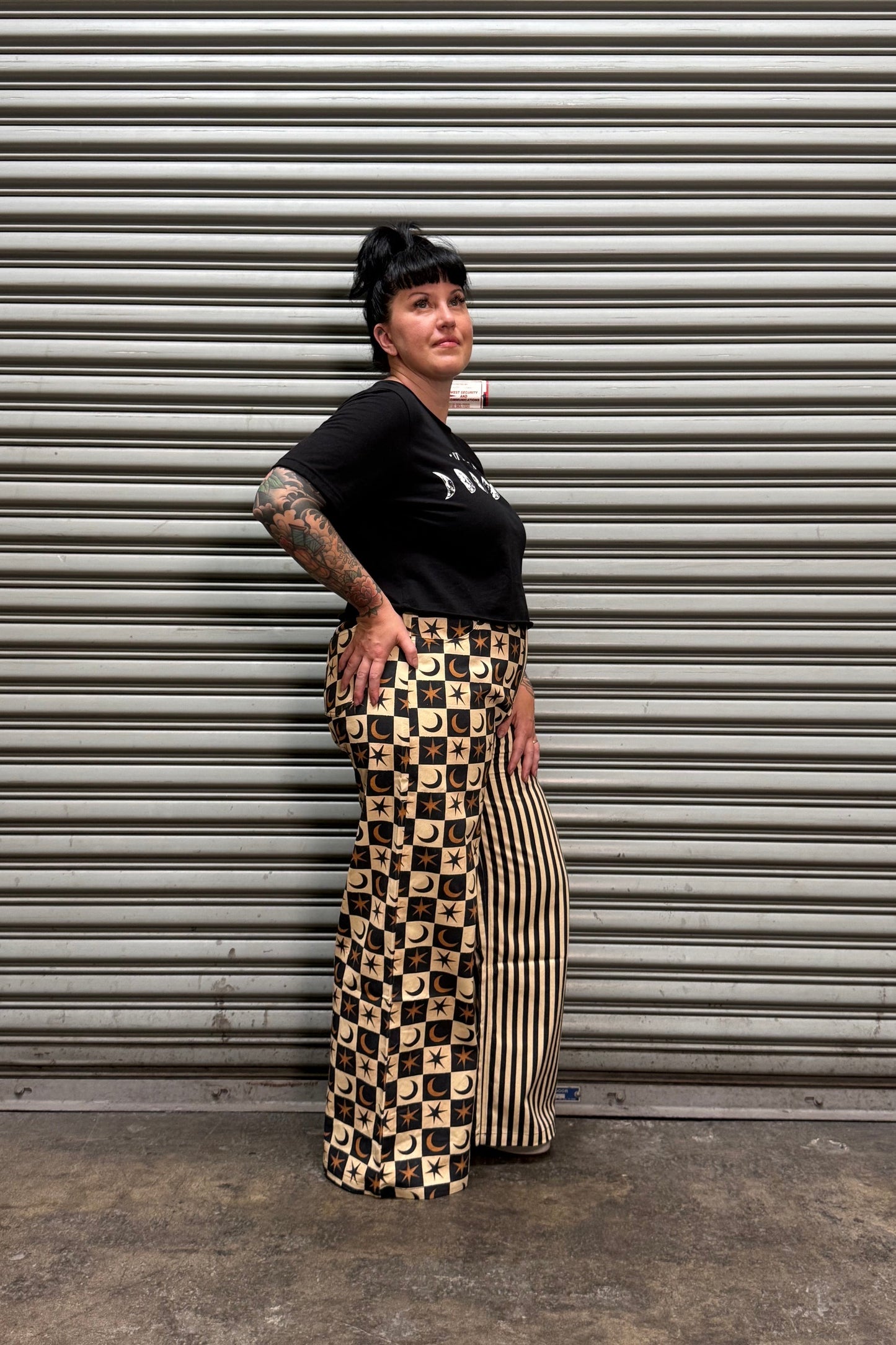 A person wearing a black t-shirt and a beige and black checkered maxi skirt, standing in front of a gray corrugated metal shutter.