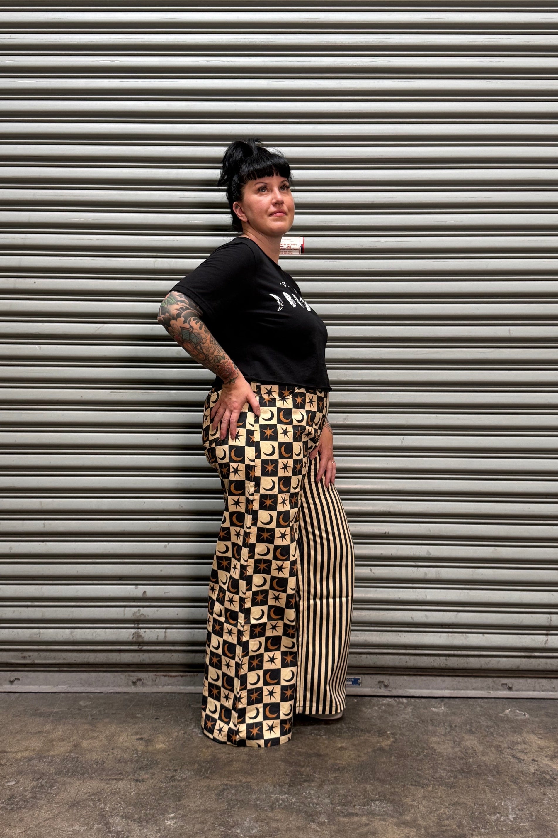 A person wearing a black t-shirt and a beige and black checkered maxi skirt, standing in front of a gray corrugated metal shutter.