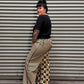 A person standing on their side against a gray corrugated metal shutter, wearing a black short-sleeve top and wide-leg beige pants with black and beige checkerboard side panels.