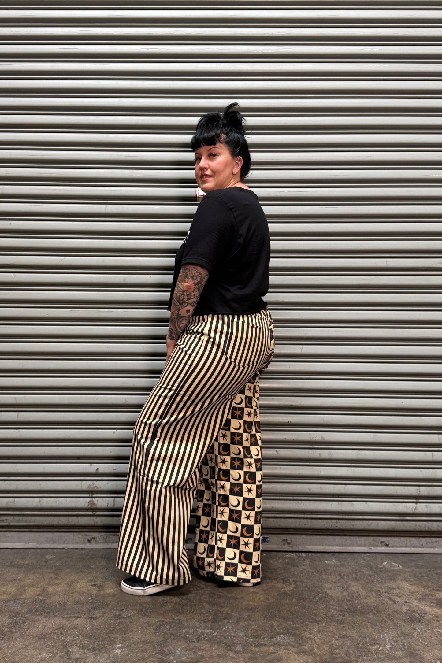 A person standing on their side against a gray corrugated metal shutter, wearing a black short-sleeve top and wide-leg beige pants with black and beige checkerboard side panels.