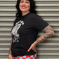 Person wearing a black t-shirt with graphic design and a red and blue checkered skirt, standing against a metallic shutter.